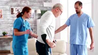 Old man with crutches helpd by young male doctor and woman