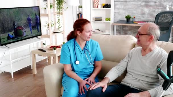 Медсестра-сиделка держит старика за руку, пока они сидят на диване. — стоковое видео