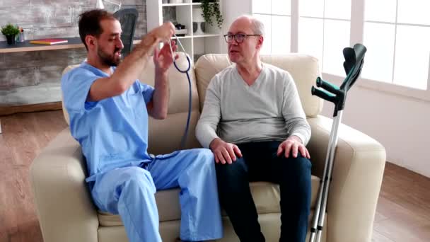 Медсестра мужчина, использующий стетоскоп, чтобы проверить сердцебиение старика — стоковое видео