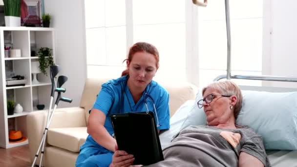 Vieille femme couchée au lit apprenant du médecin féminin à utiliser un ordinateur tablette — Video