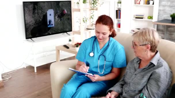 Медсестра в доме престарелых делает заметки на планшете во время разговора с пожилой женщиной на пенсии — стоковое видео