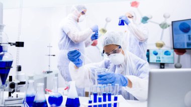 Üç bilimadamı mavi sıvıyla birkaç konteynırı inceliyor.