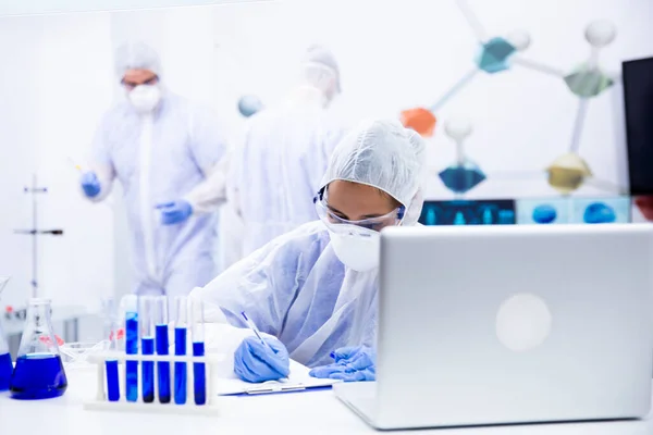 Researcher notes something on the sheet while she has several tubes and a laptop in front of her