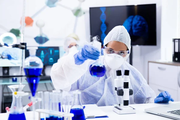 Female scientist in protection equipment holding and looking at a test tube with blue solution — Stock Photo, Image