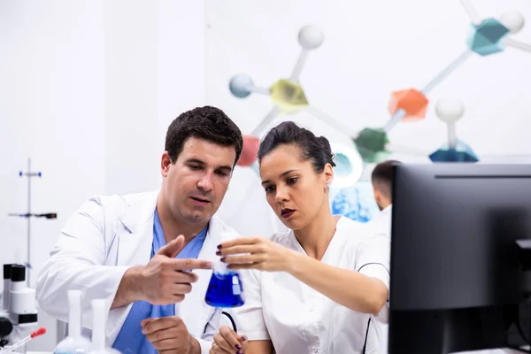 Scienziato in camice bianco e il suo assitente guardando la bottiglia con liquido blu — Foto Stock