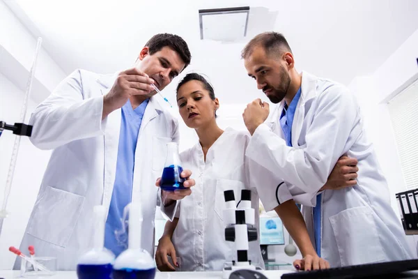 Equipo de científicos en bata blanca mirando una muestra de líquido azul humeante — Foto de Stock