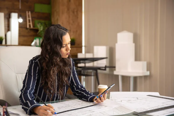 Junge Architektin schaut aufs Telefon und arbeitet an einem Projekt — Stockfoto