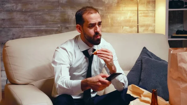 Empresario con corbata comiendo una hamburguesa mientras ve la televisión —  Fotos de Stock