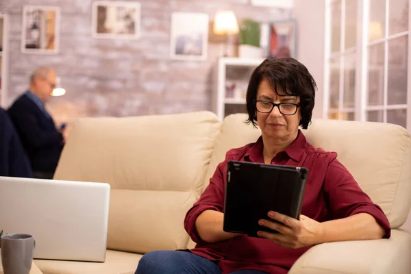 Oude oudere vrouw zittend op de bank en gebruikmakend van een digitale tablet Pc in gezellige woonkamer — Stockfoto