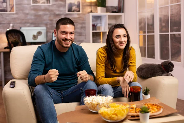 Šťastný pár jásající pro jejich oblíbený tým při sledování televize v obývacím pokoji — Stock fotografie