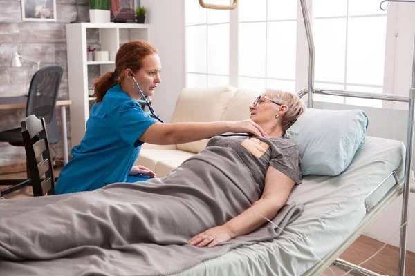 Genç bayan doktor yatakta yatan yaşlı kadının kalp atışlarını dinliyor. — Stok fotoğraf