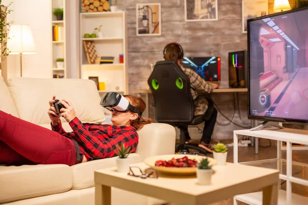 Nő fekszik a kanapén a nappaliban videojátékokat játszik egy virtuális valóság headset — Stock Fotó