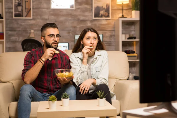 Casal comendo batatas fritas e olhando chocado na tv enquanto assiste a um filme — Fotografia de Stock
