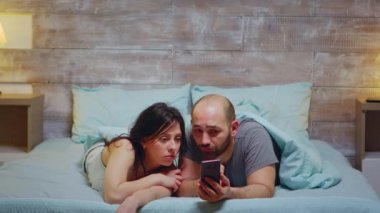 Karı koca yatakta yatarken telefonla konuşuyor.
