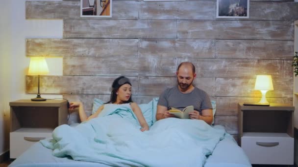 Young man reading a book while his wife is turning the lights off — Stock Video