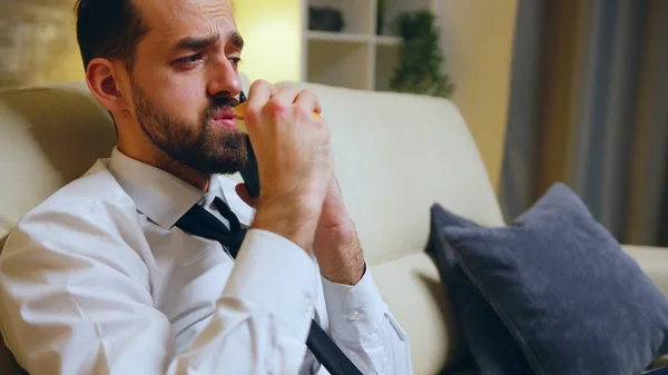 Un exitoso hombre de negocios comiendo una hamburguesa sentado en el sofá después de un día agotador — Foto de Stock