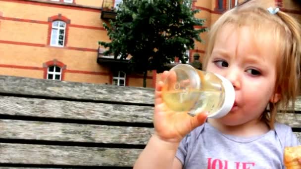 Χαριτωμένο μικρό ξανθό κορίτσι που κάθεται σε ένα παγκάκι και το πόσιμο νερό — Αρχείο Βίντεο