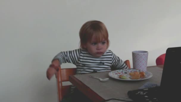 Pequeña chica rubia linda de dos años tiene cena y ver una historieta al mismo tiempo — Vídeos de Stock