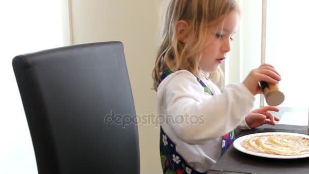 Kislány fehér ruhában egy asztalnál ül, a fahéjas palacsintát evés — Stock videók