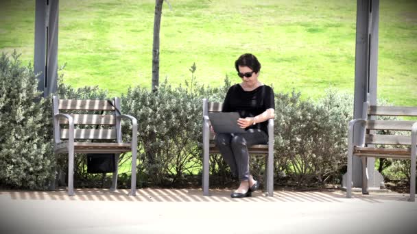 Зрелая женщина сидит на скамейке и использует свой планшетный компьютер в городском парке в солнечный летний день . — стоковое видео