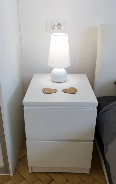 Modern white bedside nightstand with table lamp and two heart shaped cup holders. Wooden bedside cabinet with two drawers.