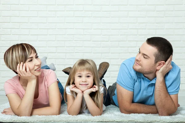 Portrait of baby and her parents lying on carpet in living room. White brick wall background, space for text. — Stock Photo, Image