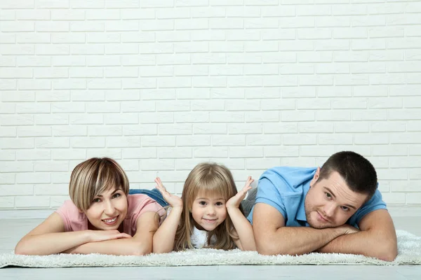 Portrait of baby and her parents lying on carpet in living room. White brick wall background, space for text — Stock Photo, Image