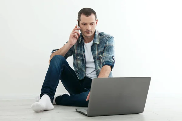 Jovem homem bonito relaxando no chão e usando laptop, telefone inteligente em casa . — Fotografia de Stock
