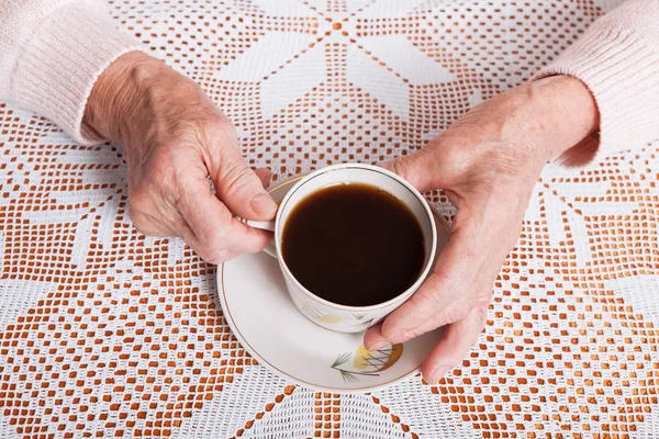 Una anciana bebe té en casa. Mujer mayor sosteniendo la taza de té en sus manos en primer plano de la mesa. Vista horizontal superior — Foto de Stock