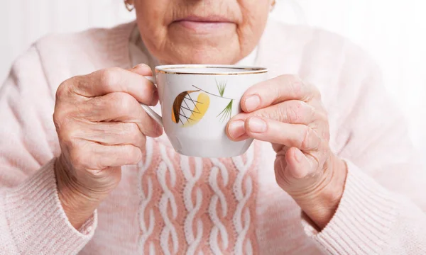 Пожилая женщина пьет чай дома. Старшая женщина держит чашку чая в руках за столом крупным планом — стоковое фото