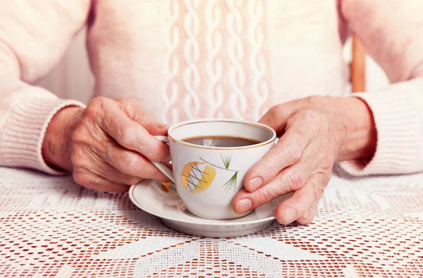Une femme âgée boit du thé à la maison. Femme âgée tenant une tasse de thé dans leurs mains à la table gros plan — Photo