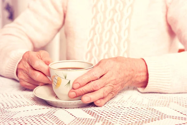 Une femme âgée boit du thé à la maison. Femme âgée tenant une tasse de thé dans leurs mains à la table gros plan — Photo