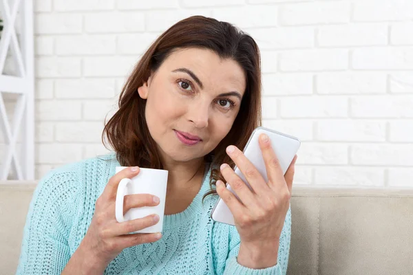 İletişim kavramı. Seyir onun akıllı telefon ve evde kanepede otururken gülümseyen çekici kadın. Orta yaşlı kadınla güzel bir kahve, sakin ol. — Stok fotoğraf