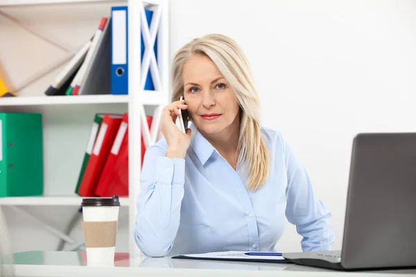 Naslouchat požadavkům klientů. Krásná žena středního věku mluvící smart telefonu a usmívá se na její pracovní místo umístění v kanceláři. — Stock fotografie