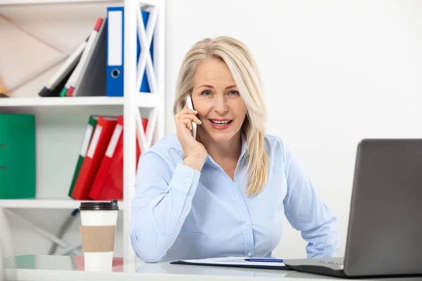 Mendengarkan persyaratan klien. Wanita paruh baya yang cantik berbicara di telepon pintar dan tersenyum di tempat kerjanya duduk di kantor . — Stok Foto