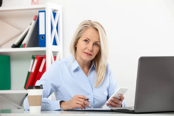 Listening to clients requirements. Beautiful middle aged woman talking on smart phone and smiling at her working place siting in the office. — Stock Photo, Image