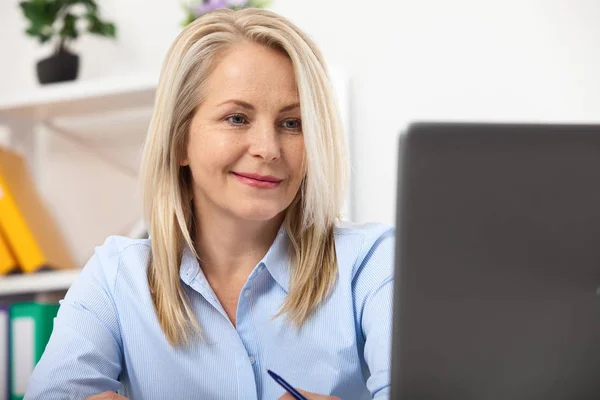 Pracę nad nowym projektem. Biznes kobieta pracuje w biurze z dokumentów. Piękna kobieta w średnim wieku patrząc na laptopa z uśmiech, podczas gdy rozmieszczenie w biurze. — Zdjęcie stockowe