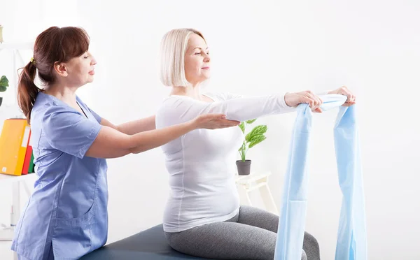 Fisioterapeuta e mulher sentados em uma cama exercitando-se com uma fita de borracha — Fotografia de Stock