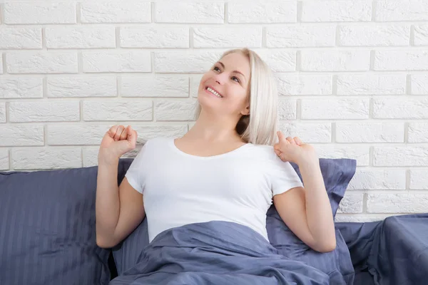 Чувственная улыбающаяся женщина с светлыми волосами сидит и растягивается в постели — стоковое фото