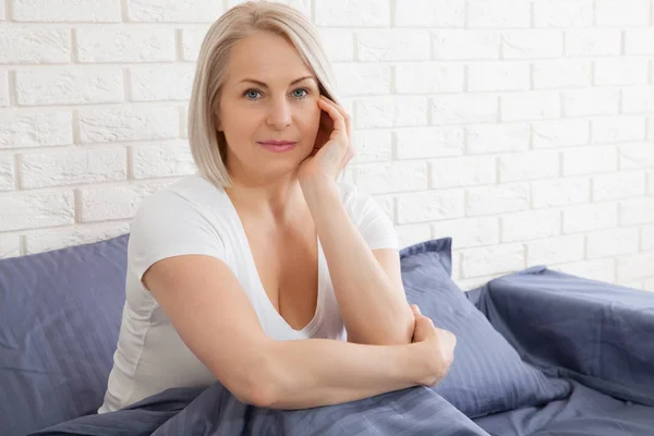Positive Frau mit Kopfkissen sitzt im Bett und lächelt zu Hause in die Kamera — Stockfoto