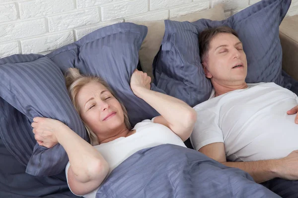 Chrapanie. Para w łóżku, mężczyzna chrapie, a kobieta nie może spać, zasłaniając uszy poduszką na chrapanie. Bliski wiek para w łóżku w domu. — Zdjęcie stockowe