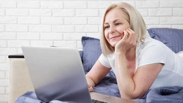 Mujer atractiva sonriente acostada en la cama usando un ordenador portátil que se comunica en línea en casa, chica feliz escribiendo en la computadora, disfrutando de las compras por Internet o charlando en las redes sociales en el dormitorio . — Foto de Stock