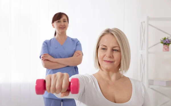 A physiotherapist helps an older woman recover from an injury through exercise with dumbbells. — Stockfoto