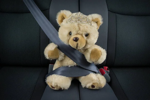 Teddy bear is sitting in a car with a tiara and is fastened with a seat belt