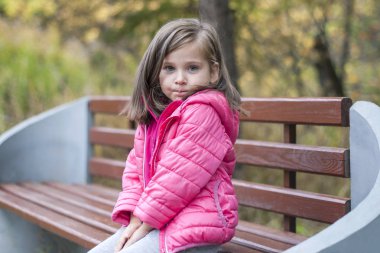 Güzel kız parkta ahşap bir bankta oturmuş sonbaharda pembe bir ceket. Duygusal portre. Çocukluk kavramı. Beyaz. Esmer uzun saçlı çocuk. Eğlence, dinlenme, yaşam biçimi