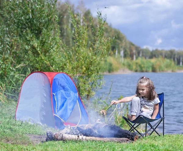 Little girl sitting near campfire. Girl roasting bread over campfire. Tourist relaxing near campfire. Rest at the lake