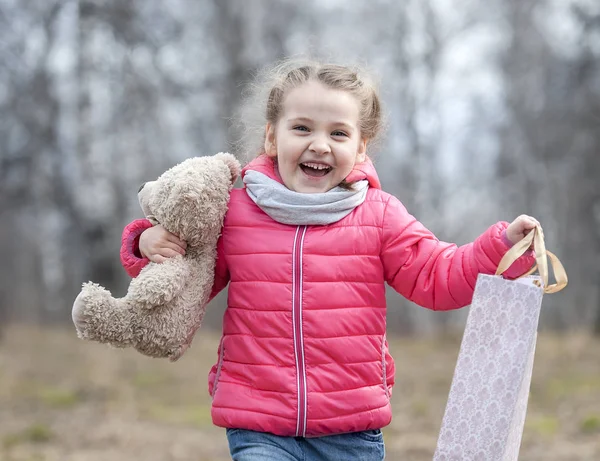 Очаровательная молодая девушка с радостью держит в руках упакованную коробку с подарком. Ребенок держит плюшевого мишку в руке на фоне дикой природы в весеннюю теплую погоду на прогулке в лесном парке — стоковое фото