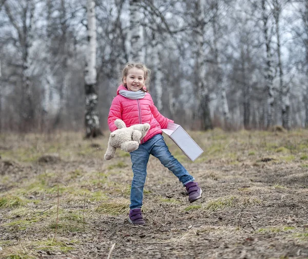 那个迷人的年轻女孩快乐地握在她的手中一个包装盒与礼物。婴儿手捧着一只泰迪熊在大自然的背景下春季暖天气在森林公园散步 — 图库照片