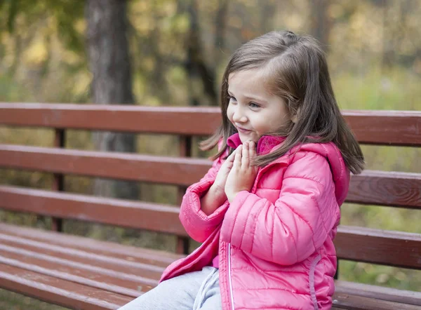 Petite jolie fille en manteau rose assise sur un banc de bois au parc en automne. Portrait émotionnel. Concept d'enfance. Caucasien. Enfant aux cheveux longs bruns. Loisirs, détente, mode de vie — Photo