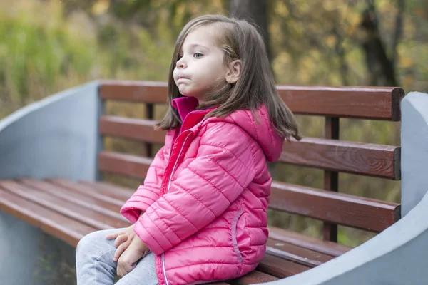 Kleines hübsches Mädchen im rosa Mantel, das im Herbst auf einer Holzbank im Park sitzt. Emotionales Porträt. Kindheitskonzept. Kaukasisch. Kind mit brünetten langen Haaren. Freizeit, Entspannung, Lebensstil — Stockfoto